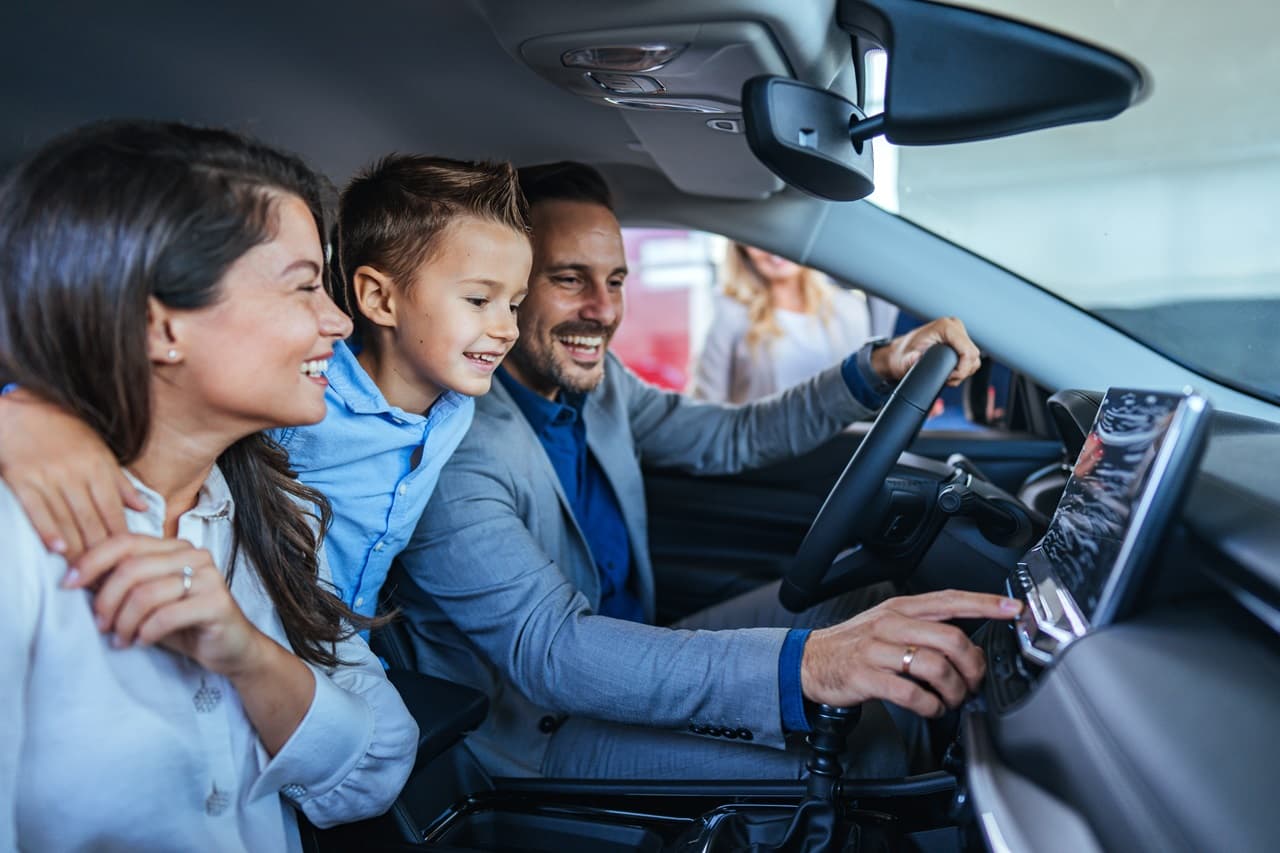 Três pessoas sentadas dentro de um veículo observam a central multimídia no painel. O interior do automóvel é moderno, com tela digital e acabamento escuro.