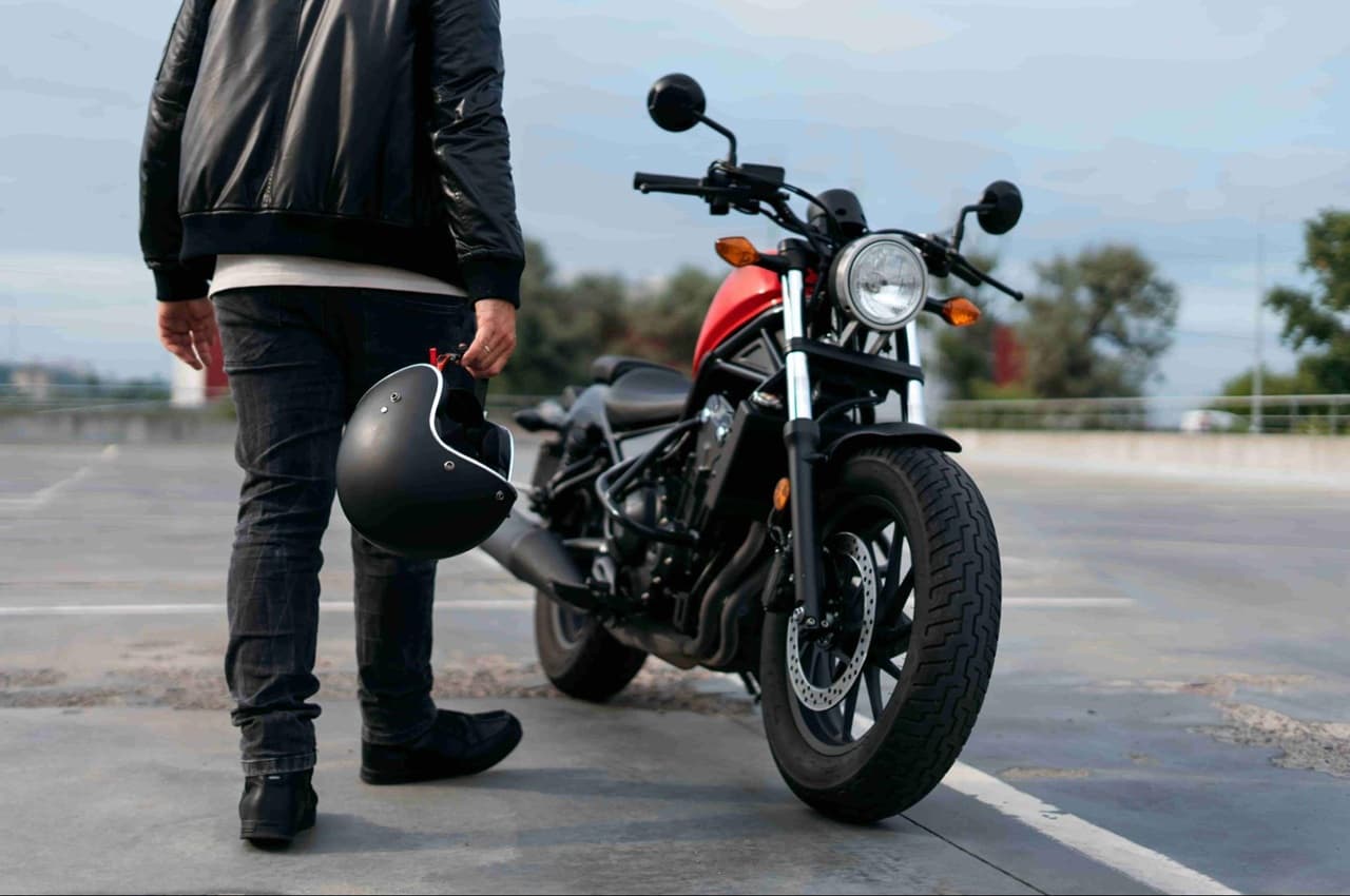 Pessoa de pé ao lado de uma motocicleta, segurando um capacete na mão, na área de um estacionamento ao ar livre.