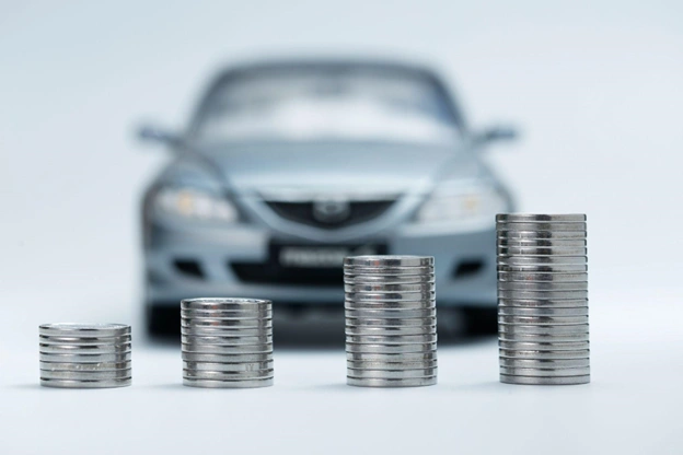 miniatura de carro prateado ao fundo com quatro pilhas de moedas em ordem crescente à frente, representando a valorização e o aumento no valor do veículo medido pelo IBV Auto. 
