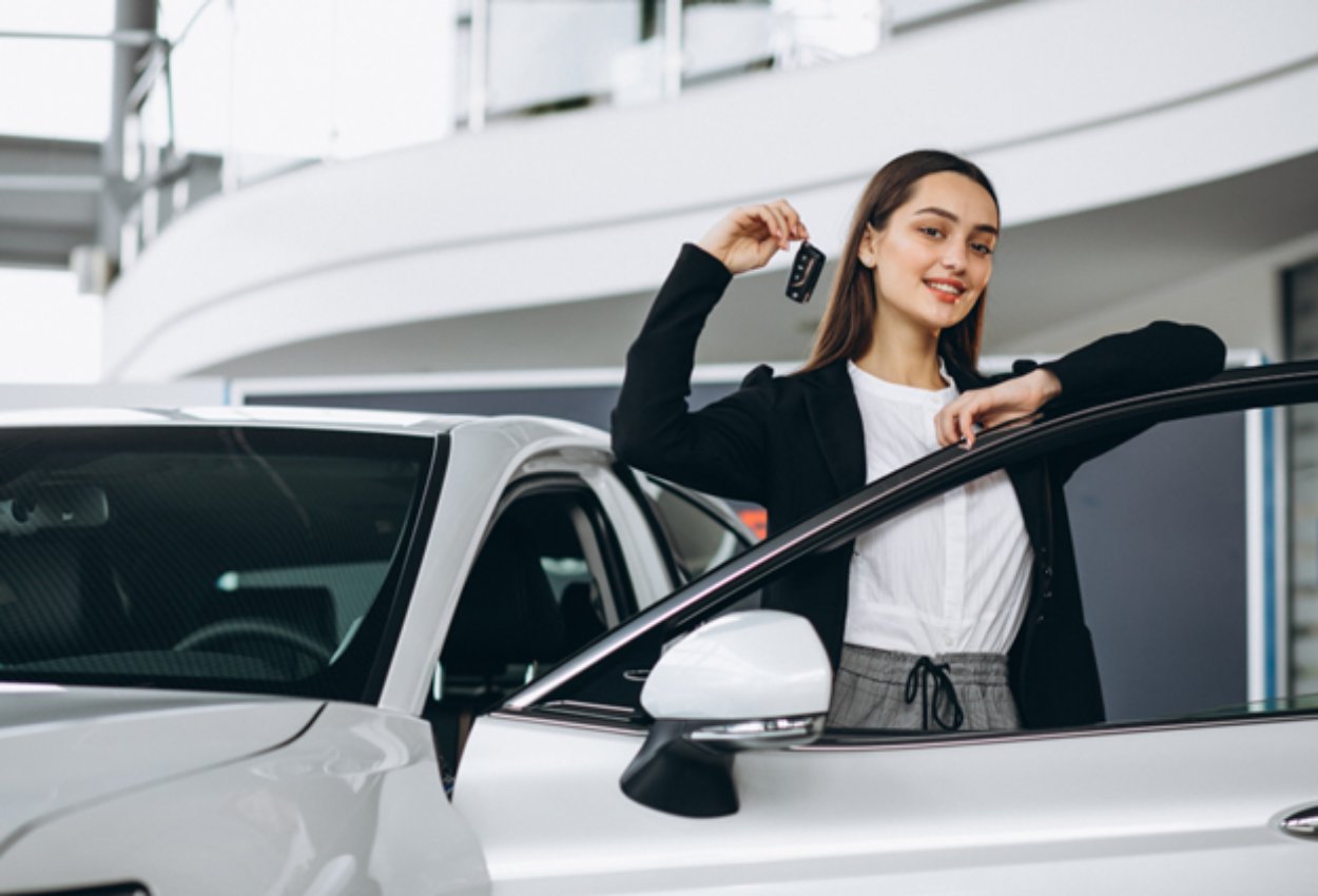 Uma mulher jovem e sorridente, vestida com um blazer preto, apoia-se na porta aberta de um carro branco em uma concessionária, enquanto segura as chaves do veículo usado.