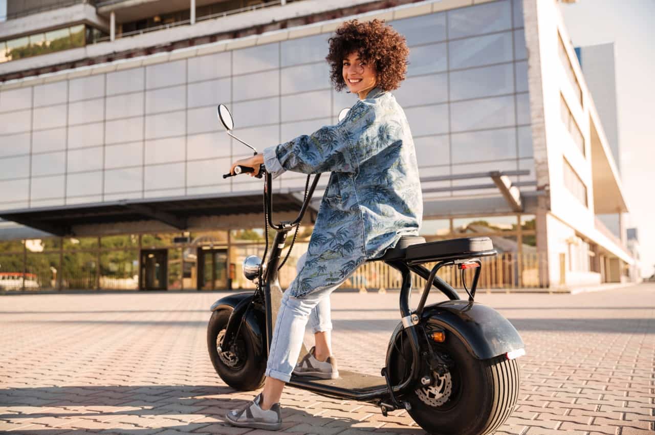 Jovem sorridente em uma moto elétrica, ao ar livre, com edifícios modernos ao fundo.