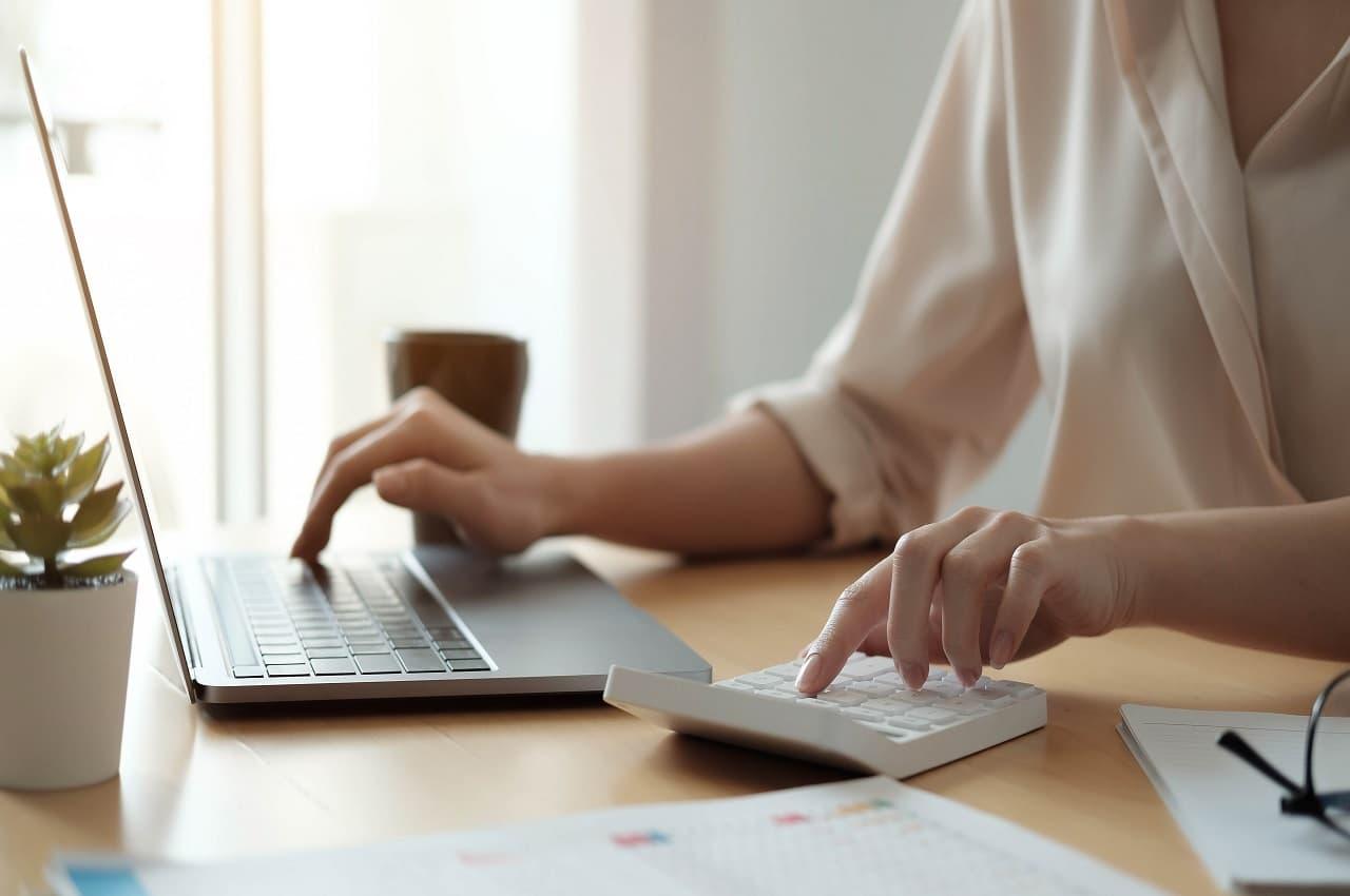 Foco em mão feminina calculando a margem de lucro na venda de carro usado com calculadora e laptop em mesa de escritório.