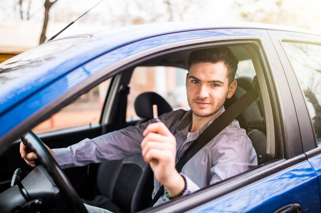 Homem com semblante feliz dentro de um carro azul. Ele está sentado no banco do motorista, olhando pelo vidro, que está aberto, e segurando a chave do automóvel.