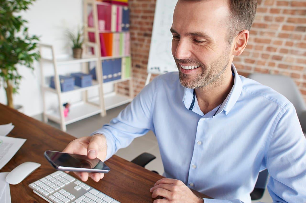 Homem sorridente sentado em uma cadeira de escritório enquanto olha para o seu celular. Ele está pesquisando sobre o novo consignado privado.