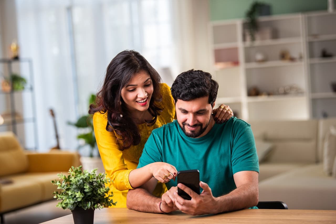 Pessoa sentada à mesa segurando um smartphone, enquanto outra pessoa ao lado aponta para a tela do aparelho. Há um vaso de planta sobre a mesa e móveis ao fundo.