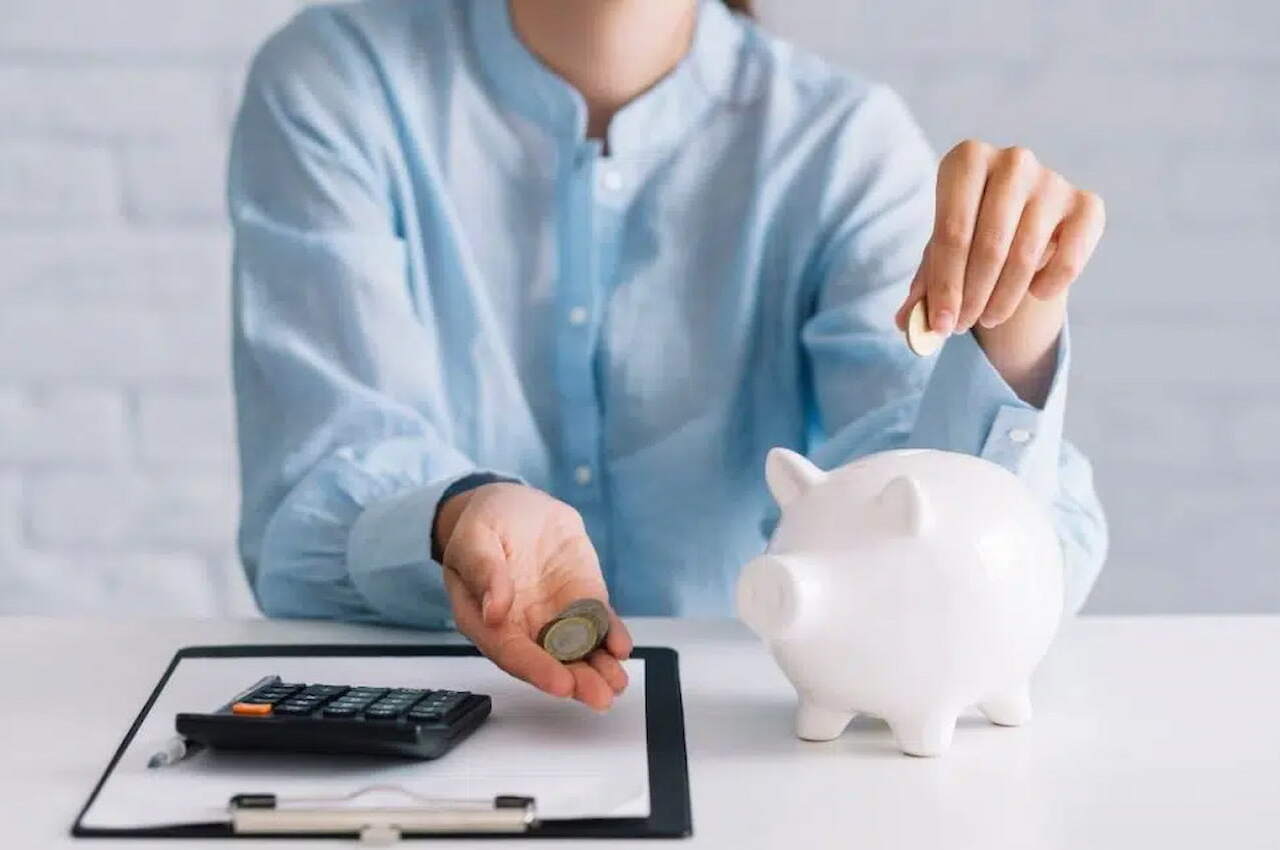 Mulher segurando moedas e colocando moeda em um cofre de porquinho, com calculadora e prancheta na mesa, simbolizando a taxa CDI. 