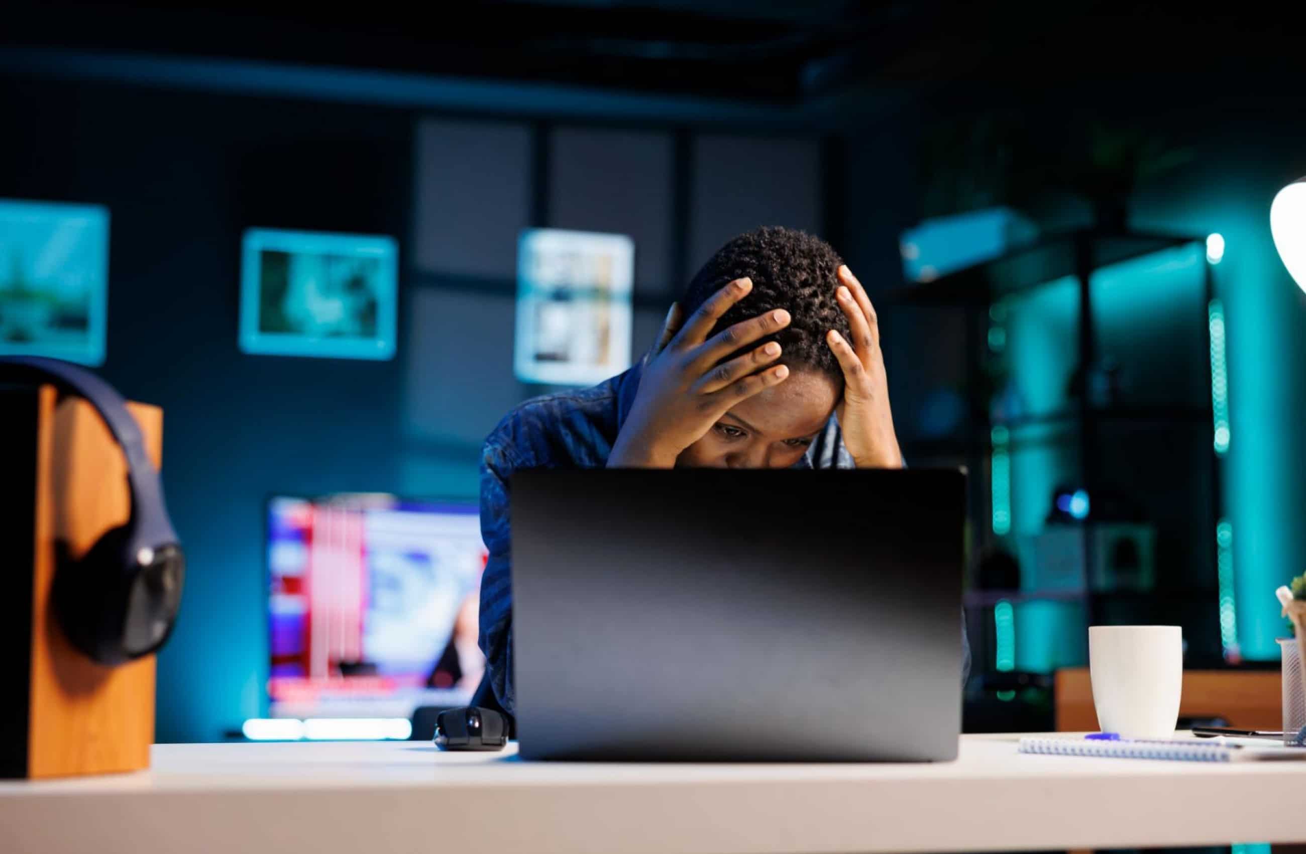 Pessoa sentada diante de um notebook, com as mãos na cabeça e expressão de preocupação, em um ambiente escuro — representando o estresse e os prejuízos causados por um golpe ou fraude digital.
