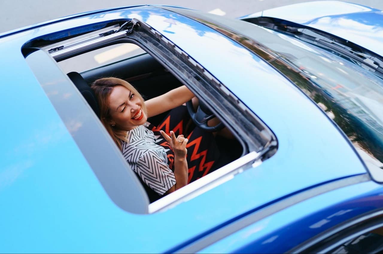 Teto solar de um veículo aberto e uma mulher sorridente em carro azul, mostrando satisfação e alegria ao dirigir.
