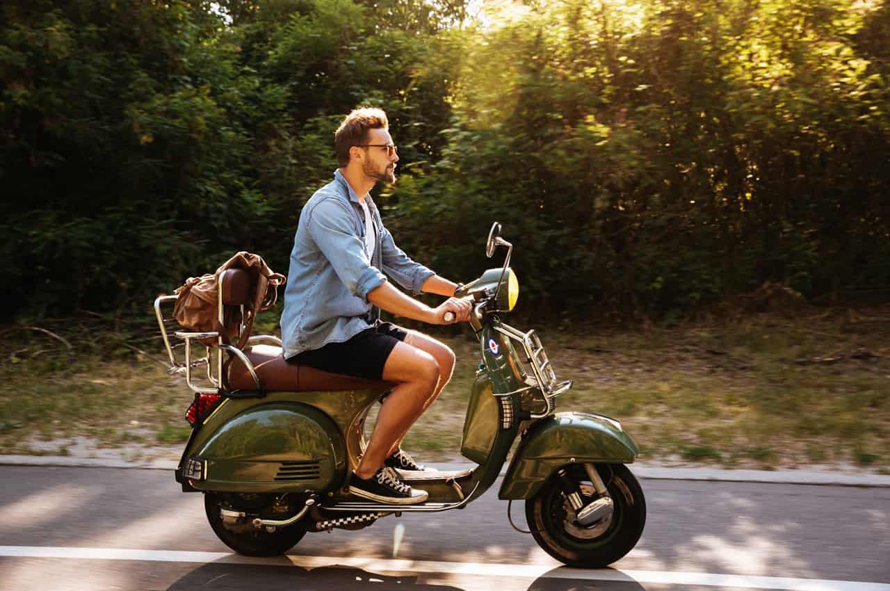 Homem de óculos escuros pilotando uma scooter clássica verde em uma estrada ao lado de uma floresta, durante o pôr do sol.