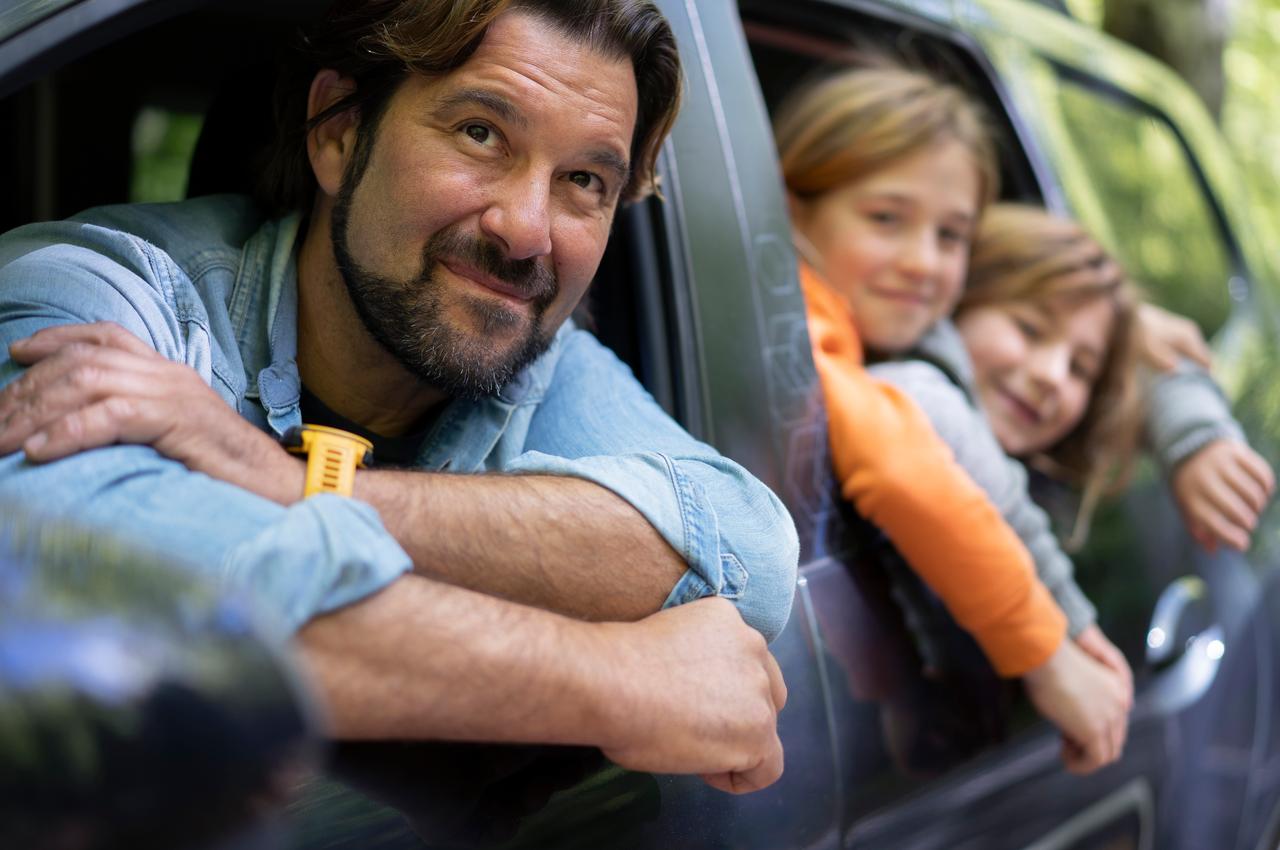 Homem sorridente com barba e duas crianças, sentado na janela do carro, curte um momento ao ar livre em família.