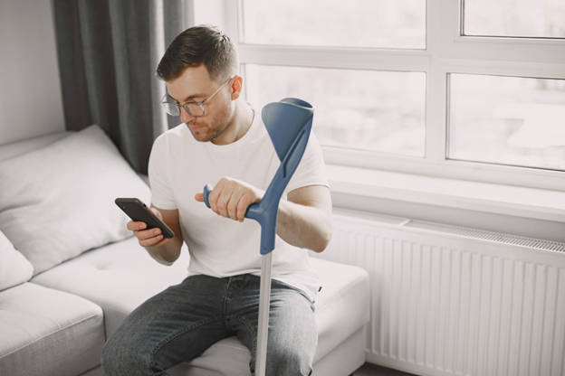 Homem sentado no sofá usando muleta e consultando o celular, ilustrando a importância do seguro de acidentes pessoais para apoio financeiro durante a recuperação.