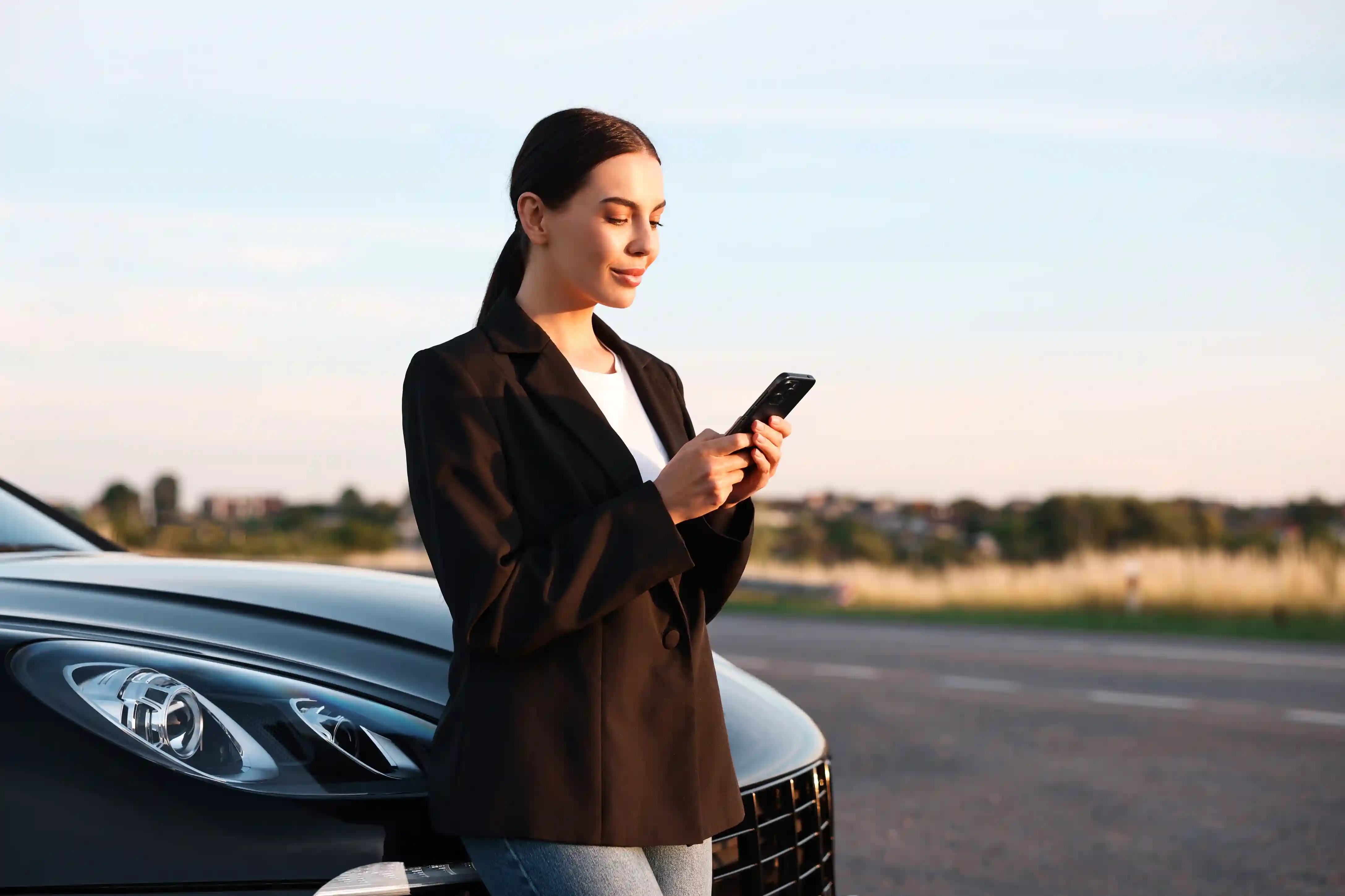 Mulher ao lado do carro consultando o celular na estrada, ilustrando a contratação e assistência do Seguro RCF-V. 
