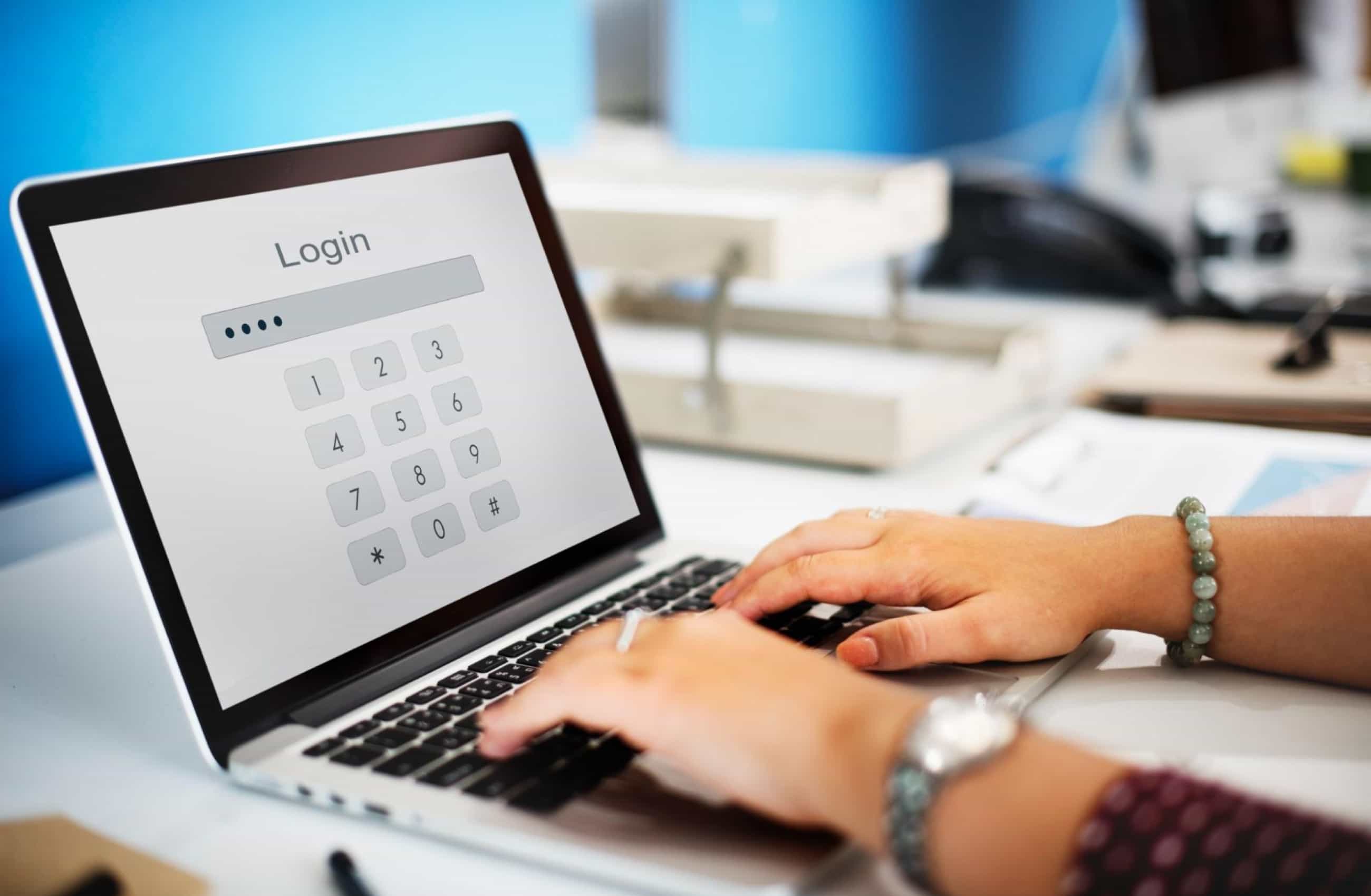 Mãos de uma pessoa digitam no teclado de um laptop que exibe uma tela branca de login com teclado numérico virtual e quatro pontos de senha. A cena representa a importância da senha forte.